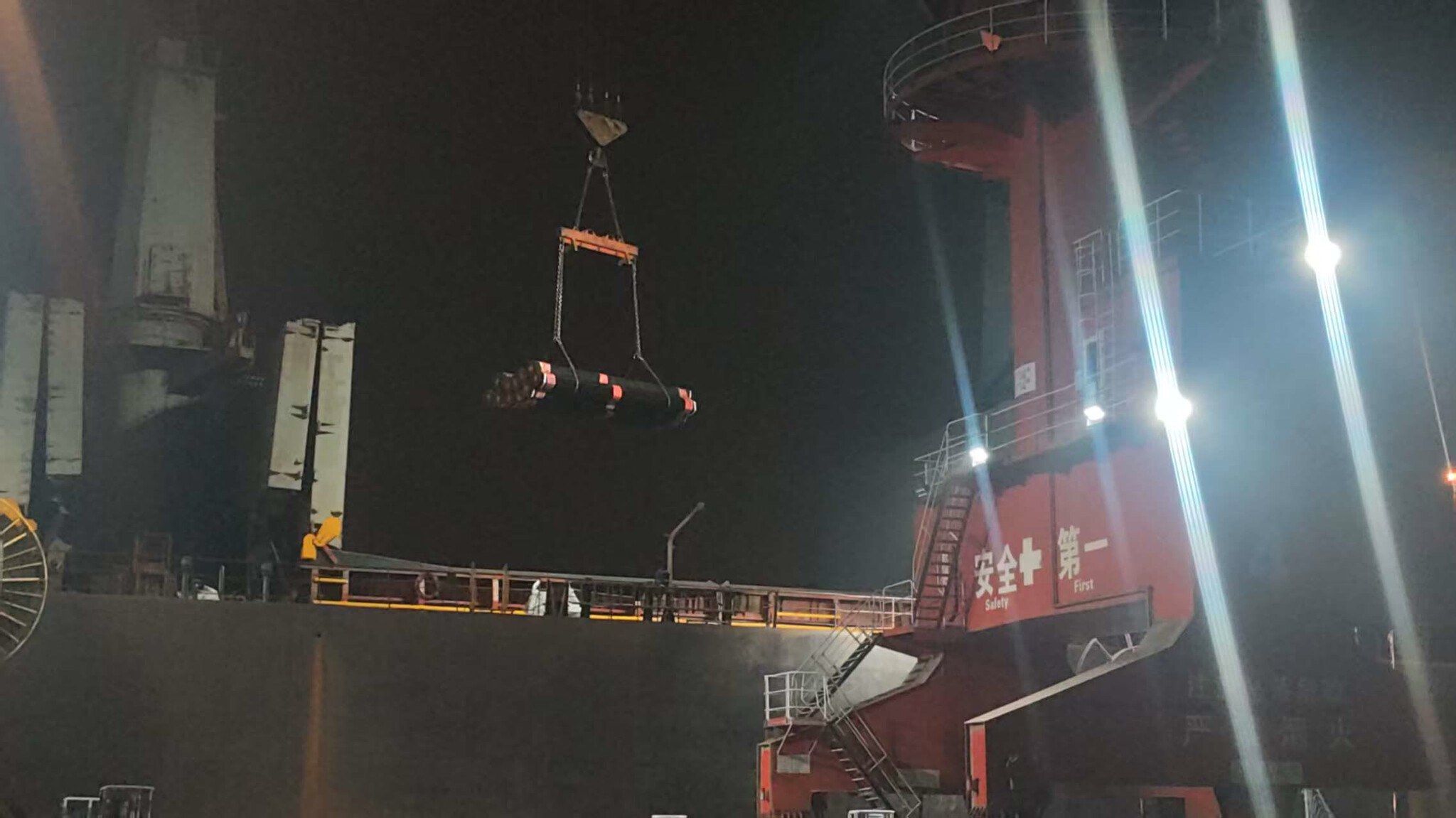 A ship’s crane lowering pipe bundles into the hold at night