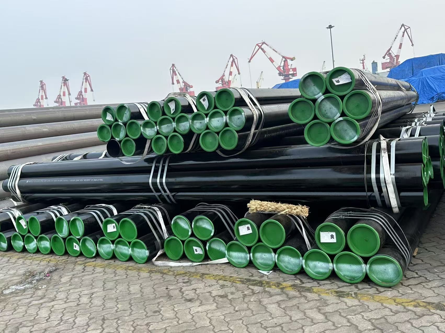 Stacked seamless pipe with green end caps on the quay, port cranes in the background