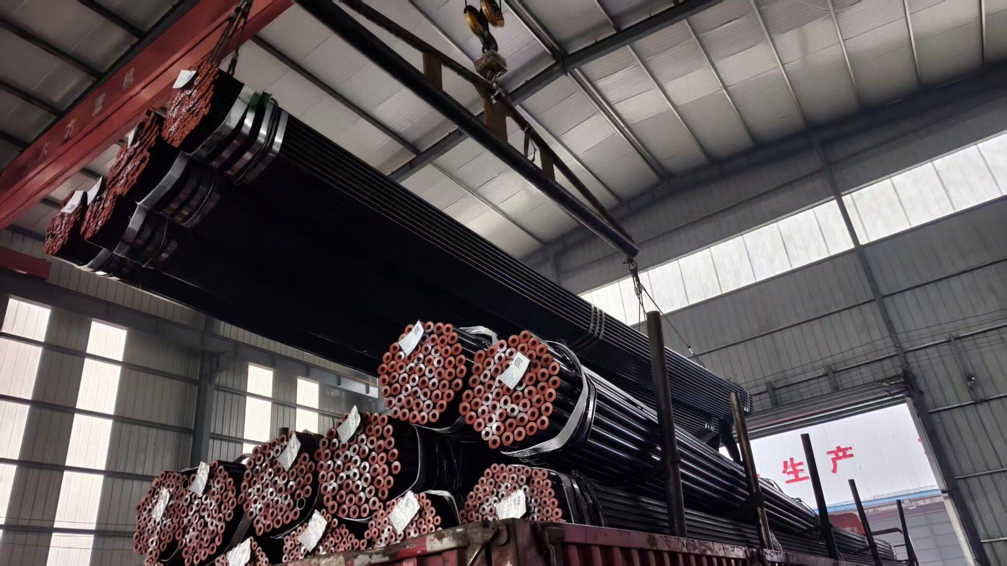 An overhead crane loading black seamless pipe bundles onto a truck inside a warehouse